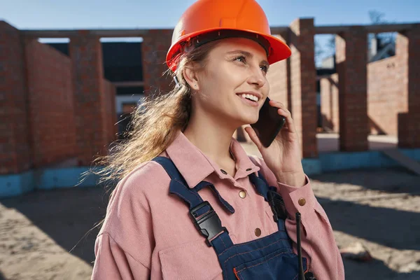 Esperançosa jovem senhora no construtor uniforme conversando no telefone — Fotografia de Stock