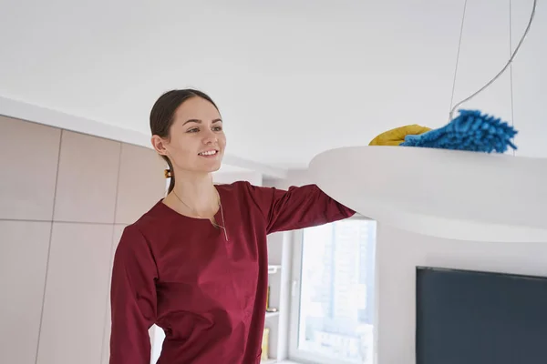 Χαμογελαστή καθαρίστρια που ξεσκονίζει το φωτιστικό οροφής — Φωτογραφία Αρχείου