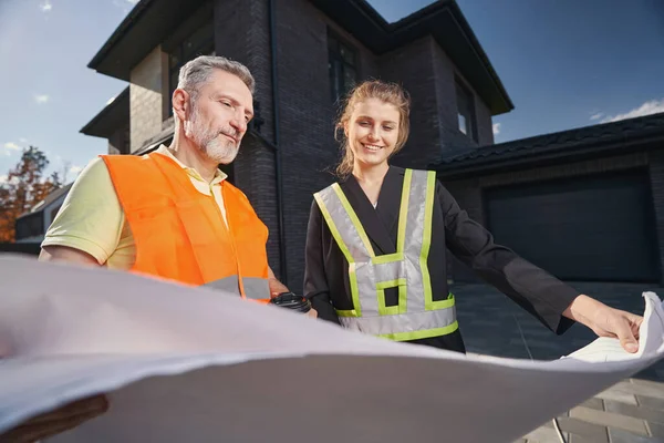 Åldrande byggmästare och kvinna i byggnadsvästar stirrar på ritningar — Stockfoto