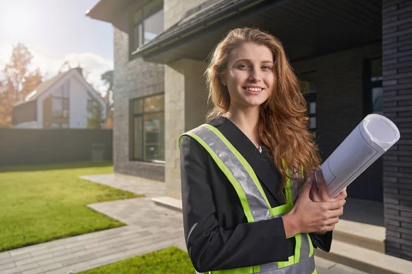 Cheerful female construction engineer with blueprints smiling to camera — Stockfoto