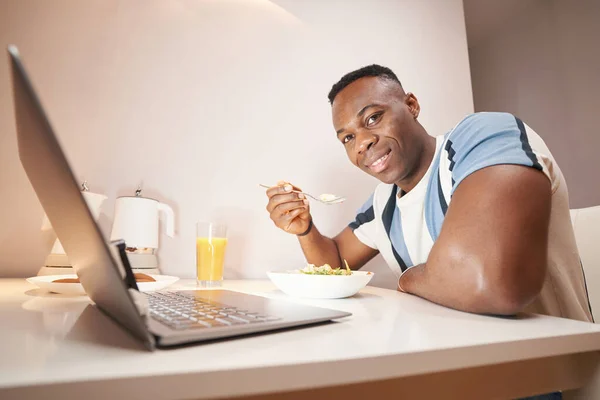 Seorang pria melihat kamera sambil makan sebelum laptop — Stok Foto