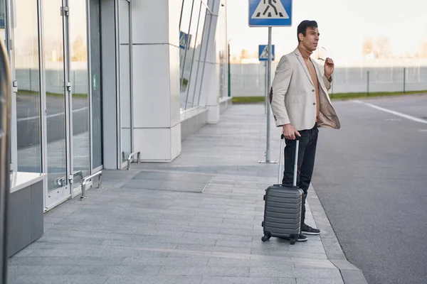 Mann nimmt Brille ab, während er mit Koffer im Freien steht — Stockfoto