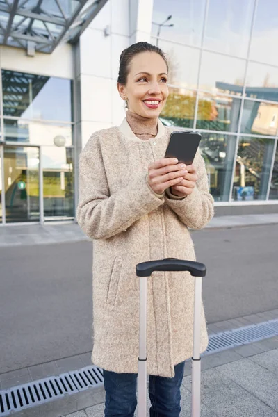 Zadowolona kobieta klikając na smartfon obok budynku lotniska — Zdjęcie stockowe