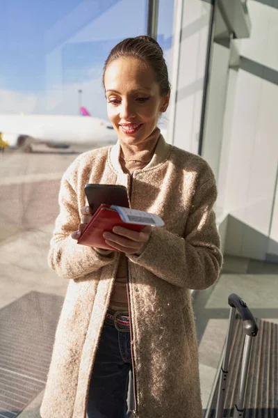 Vrolijke dame in jas met behulp van smartphone in de buurt van luchthaven venster — Stockfoto