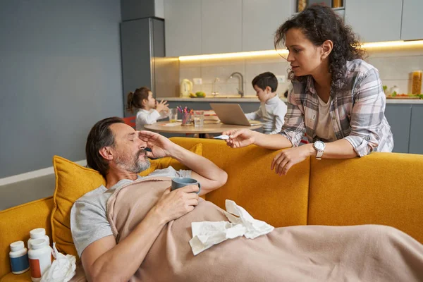 Woman speaking to man on couch while holding thermometer — Stock Photo, Image