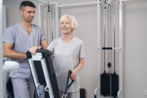 老年妇女站在康复健身房设备的躺椅前 用左手拉开缆绳 — 图库照片