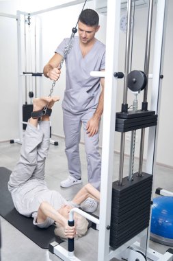 Fizyoterapist gözetiminde yaşlı bir kadın kablo makinesiyle bacak kaldırıyor.