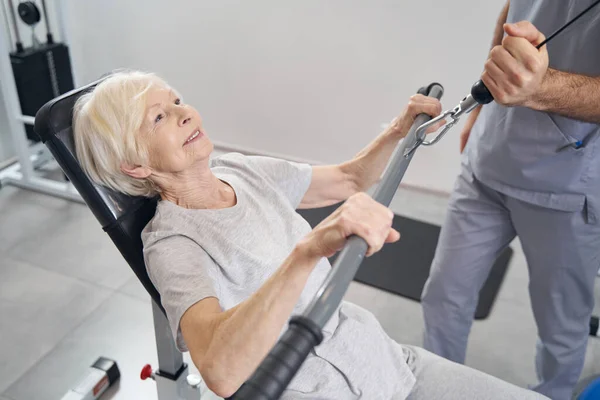 Spor aletleri koltuğunda kol egzersizi yapan yaşlı bir kadın. — Stok fotoğraf