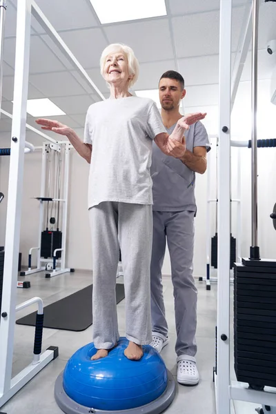 Seniorin steht mit Hilfe von Therapeut auf Fitnessball — Stockfoto