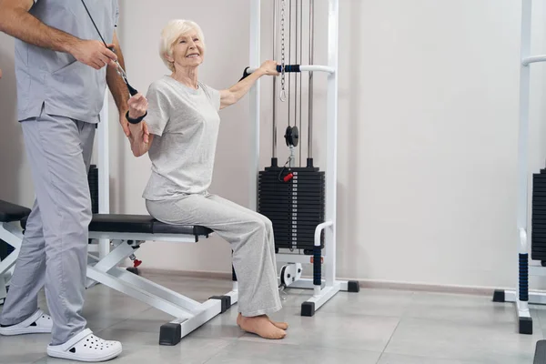 Pleased elderly woman pulling handle and cable of strength machines — Stock Photo, Image