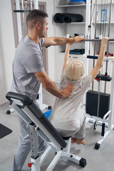Fisioterapeuta inclinado paciente anciano hacia adelante durante el ejercicio en la máquina de fuerza —  Fotos de Stock