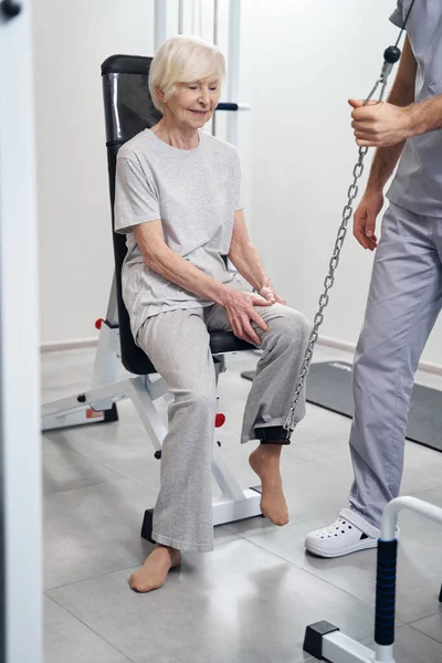 Medical specialist lifting leg of senior citizen with cable machine — Stock Photo, Image