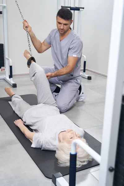 Cirurgião ortopédico cuidadoso levantando perna pensionista com máquina de cabo — Fotografia de Stock