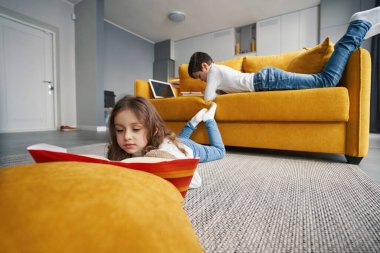 Kardeşim kanepede yatarken yerde kitap okuyan bir kız.