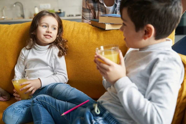 Little girl with juice glass on sofa with brother — Stock Photo, Image