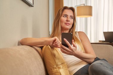 Genç bayan otel odasındaki kanepede akıllı telefon kullanıyor.