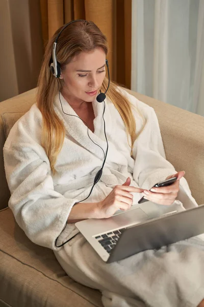 Mulher trabalhando no fone de ouvido com laptop usando smartphone — Fotografia de Stock