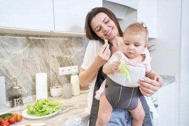 Bebek taşıyıcısında çocuğu olan zarif esmer anne elinde salata tutuyor.