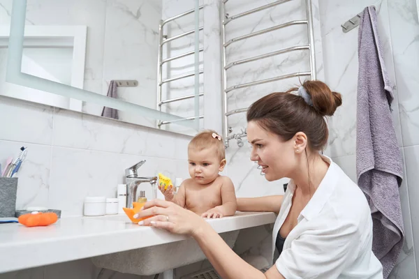 Cute little kid sitting in washbasin while playing with bath toys in room indoors — Stock Photo, Image