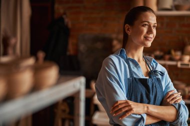 Modern sanat atölyesinde fotoğraf makinesine poz veren önlük giymiş güzel bir kadın sanatçı.