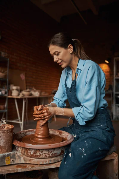 Piękne pozytywne rzemiosło kobieta noszenie fartuch podejmowania gliniany garnek na kole ceramiki w warsztacie — Zdjęcie stockowe