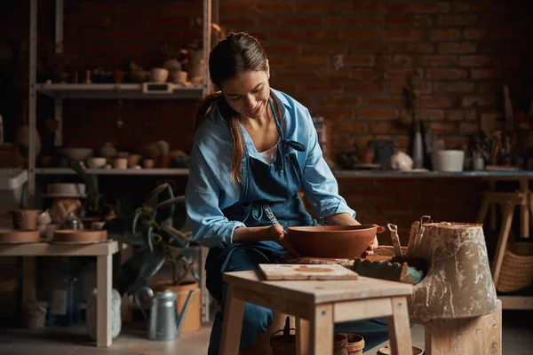 Przyjazny atrakcyjny żeński rzemieślnik noszący fartuch modelujący ceramiczne zastawy stołowe w warsztacie garncarskim — Zdjęcie stockowe