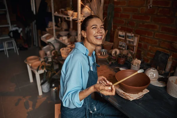 Hermosa mujer positiva con delantal sentado en el escritorio mientras pinta tazón de cerámica en taller de cerámica —  Fotos de Stock