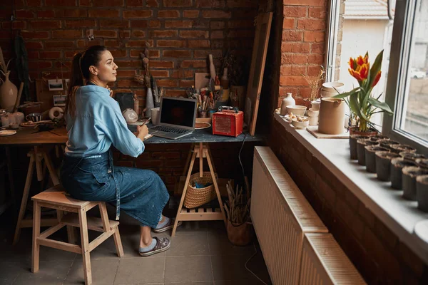 Задумчивая очаровательная леди, работающая за ноутбуком в художественной студии — стоковое фото