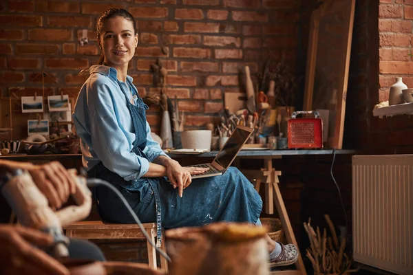 Morena mujer artesanal caucásica sentada en la silla mientras posando en la cámara de fotos en el taller de cerámica —  Fotos de Stock