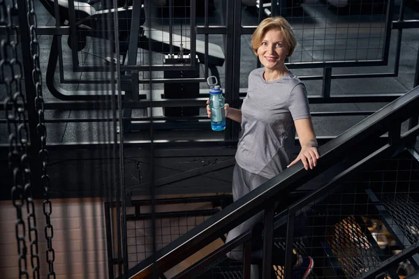 Cheerful sporty woman with drink standing on stairs in gym — Stock Photo, Image