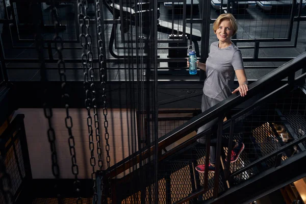 Joyful woman with drink standing on stairs in gym — Stock Photo, Image