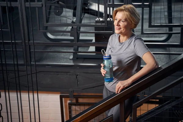 Cheerful woman with drink standing on stairs in gym — Stock Photo, Image
