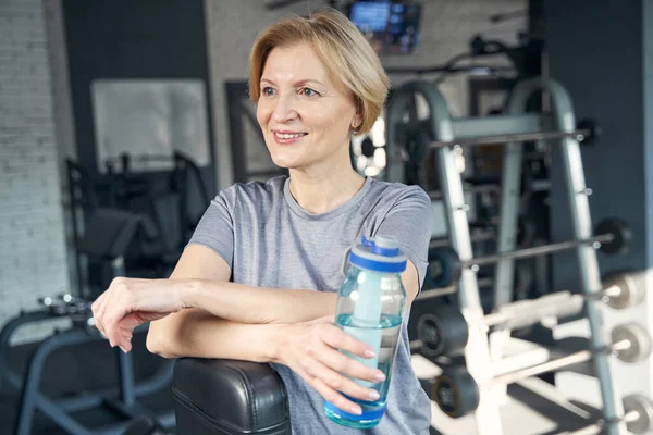 Mujer alegre con botella de agua de pie en el gimnasio — Foto de Stock