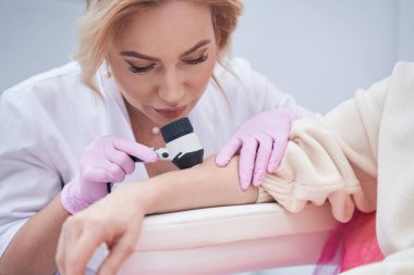 Odaklanmış doktor dermatoskopla hastanın cildini kontrol ediyor.