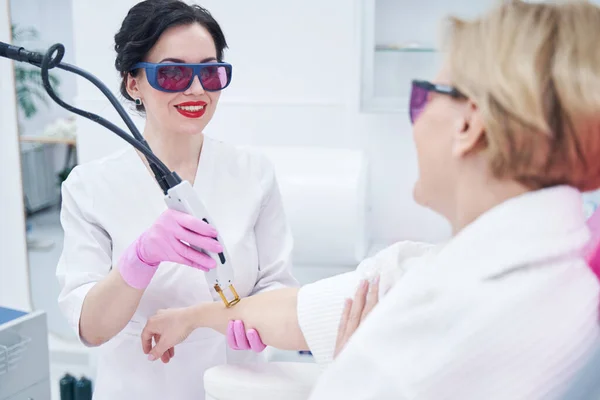 Cosmetologista alegre removendo o cabelo do braço da mulher com dispositivo laser — Fotografia de Stock