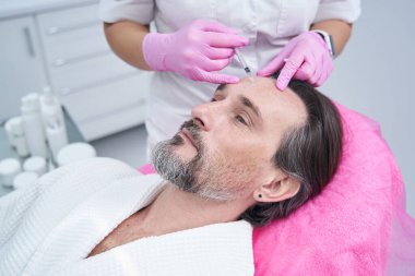 Doktor kozmetik uzmanı klinikte yaşlı bir adamın kırışık alnına dolgu yapıyor.