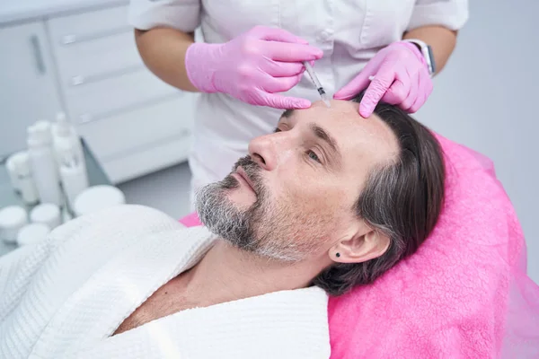 Doktor kozmetik uzmanı klinikte yaşlı bir adamın kırışık alnına dolgu yapıyor.