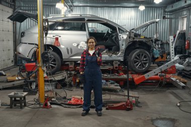 Araba tamir aletini tutan ve tamirhanedeki arabanın yanında kameraya bakan tam uzunlukta bir kadın işçi.