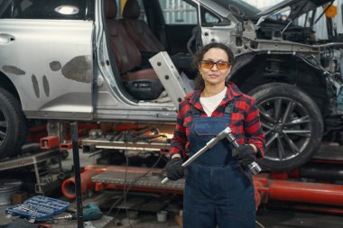 Kadın araç teknisyeni elinde ingiliz anahtarıyla garajda otomobilin yanında duruyor.