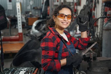 Güvenlik gözlüklü neşeli bayan çalışan kameraya bakıyor ve araç metalini tutarken gülümsüyor.