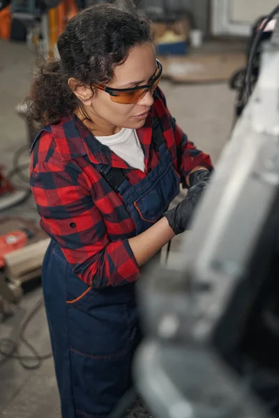 Female vehicle technician in safety glasses repairing car at auto service garage