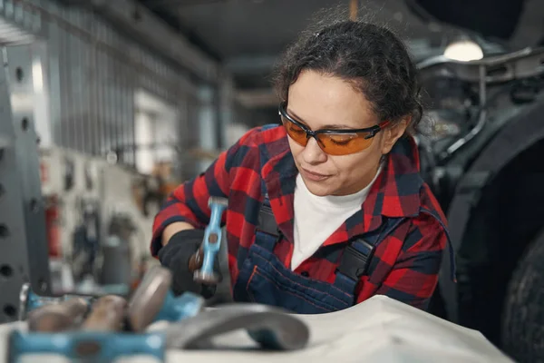 Female worker in safety glasses repairing vehicle parts with hammer in auto repair workshop
