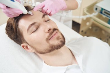 Güzellik uzmanı ultrasonik deriyi temizlerken ve alnındaki gözenekleri temizlerken adam gözleri kapalı dinleniyor.