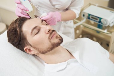 Alnında ultrasonik deri fırçasıyla güzellik salonunda deri tedavisi gören bir erkek.