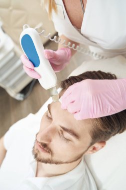 Erkek hasta için ultrasonik bir aletle alnın cildini temizleyen güzellik terapisti.