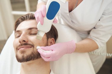 Güzellik uzmanı, erkek yüzündeki gözeneklerden ultrasonik temizleyici ile kir ve hücresel kalıntıları temizliyor.