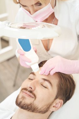 Kozmetik uzmanı, erkek hastanın alnını ultrasonik aletle ovalayıp pamukla siliyor.