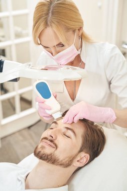 Maskeli beyaz kadın, memnun bir adamın alnını ovuyor ve ultrasonik dalgalarla temizliyor.