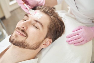 Kafasına maske takmak için krem sürerken gülümseyen beyaz bir adam.