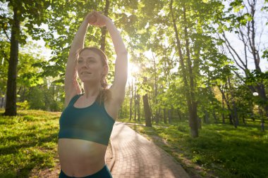 Mutlu sportif dişi, güneşli bir günde ağaçların altında parkta koşup kollarını kaldırarak esnetiyor.